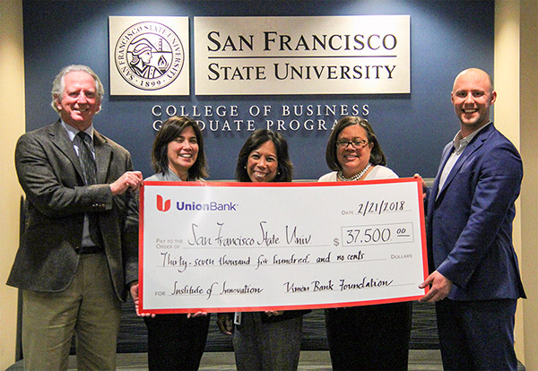 Management Professor Tom Thomas, Katerina Villanueva, Lisa Corpuz, Dean of the College of Business Linda Oubre, and Daniel Meals standing holding a blown-up facsimile of a check at the SFSU Downtown Campus