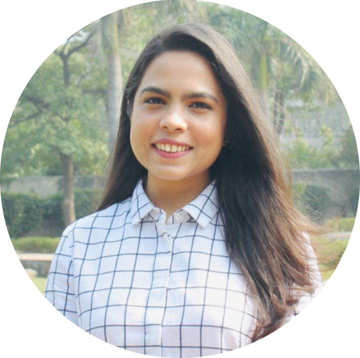 Arushi Bhambri, smiling alumna with long brown hair, wearing button-down, checkered white top. Greenery in background 