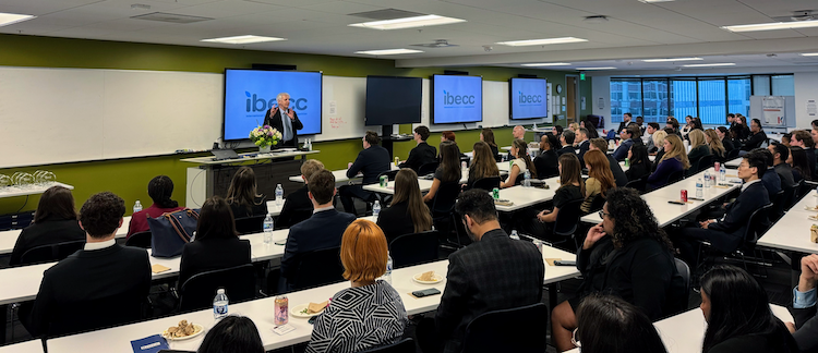 2025 International Business Ethics Case Competition at SFSU Downtown Campus, attendees seated at tables in a large meeting room and a speaker at the front of the room with monitors behind them 
