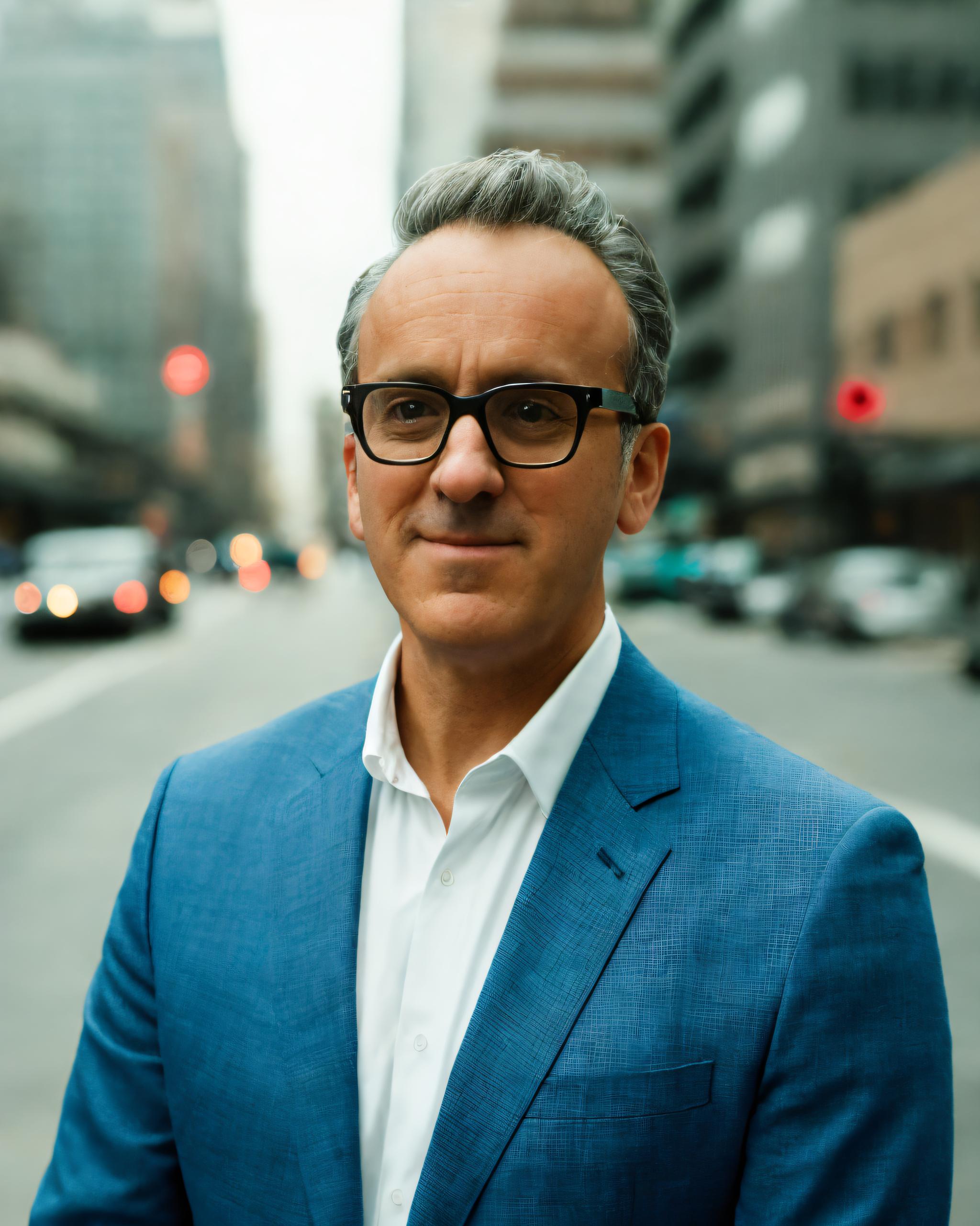 Carlos Flores, man standing on city street, wearing blue blazer, white button-down shirt and dark-rimmed glasses