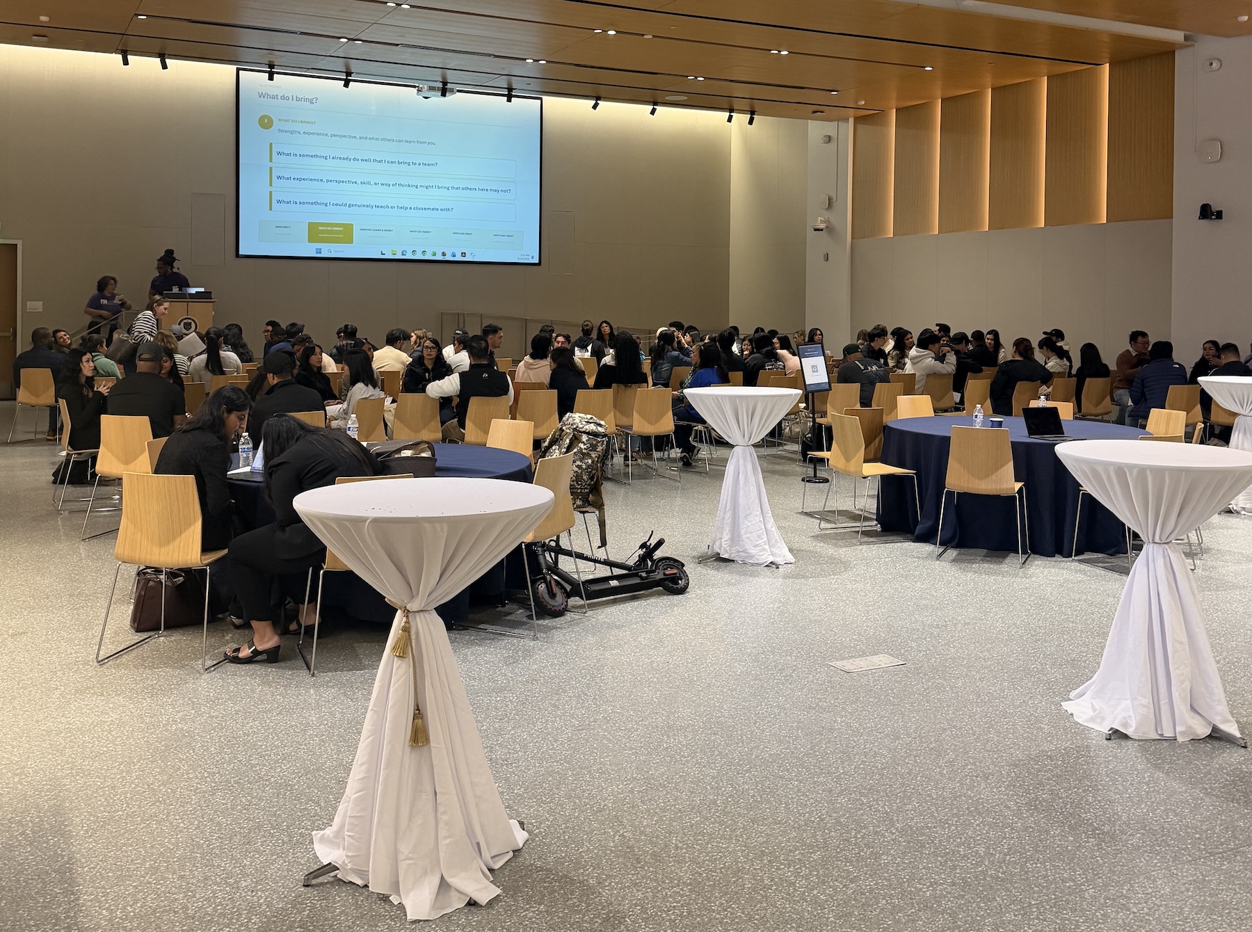 Wood-paneled, modern auditorium/reception room full of people sitting watching a presentation on a large monitor, with cocktail tables in the forefront