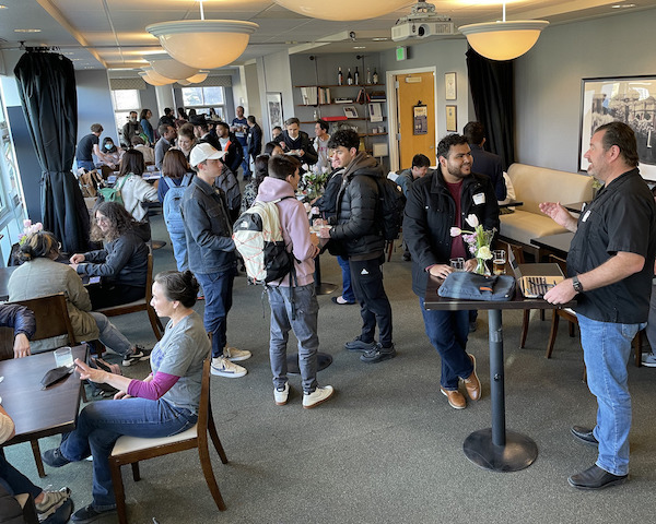 Group of students, faculty gather with around tables, chairs in Vista Room restaurant for Demo Days event in Februrary 2023