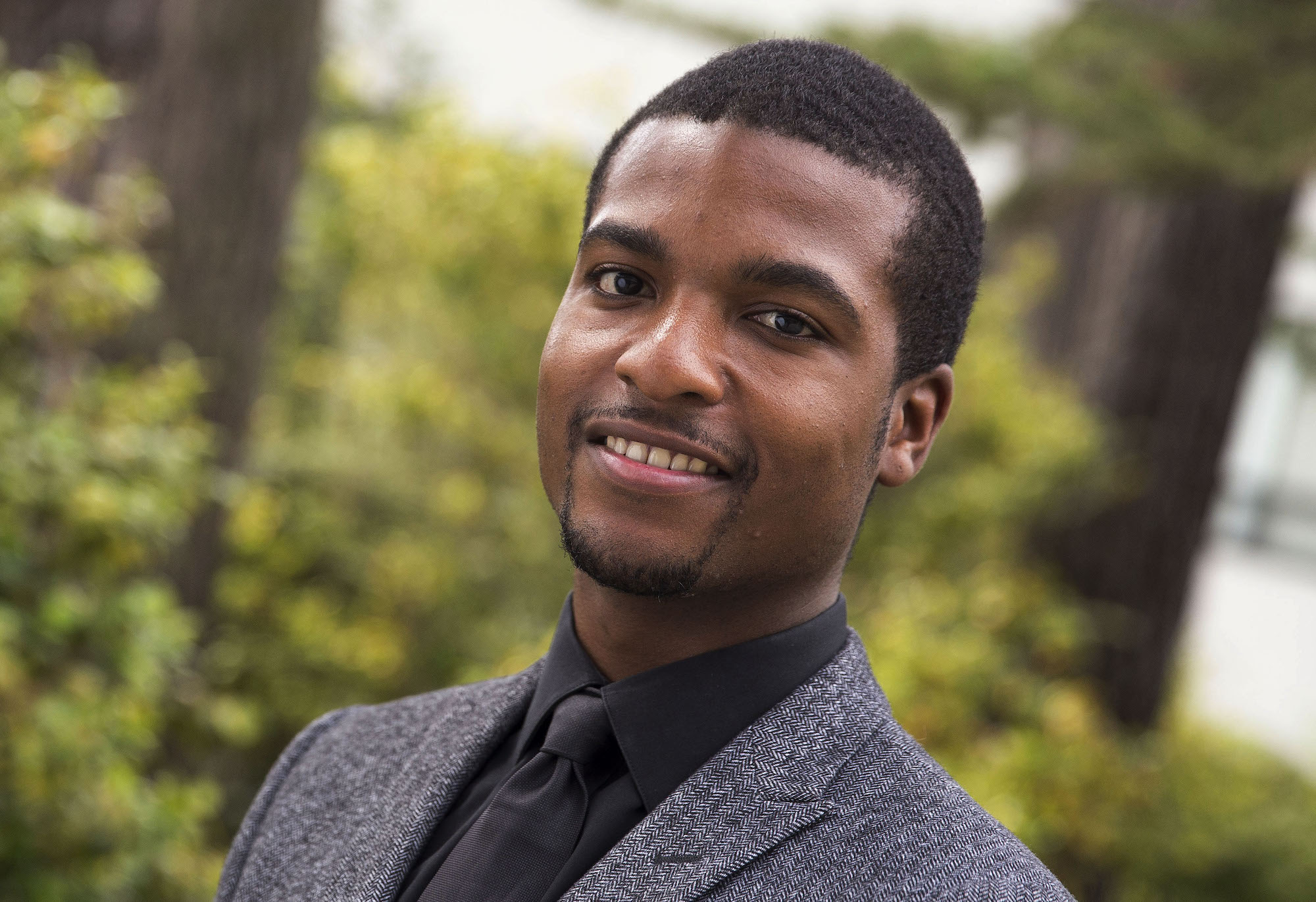 male student dressed in suit jacket and tie, standing outside with trees in background