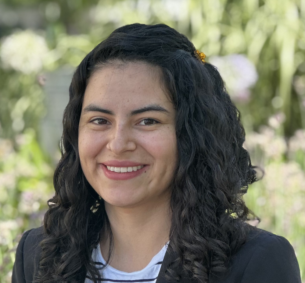 Lina Perez, headshot of smiling female student with dark long hair, wearing a dark blazer and white top.  Greenery in background.