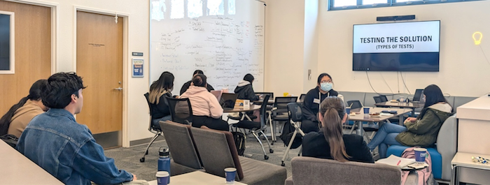 Students sitting in a room listening to a speaker standing at the front of the room in from on a large screen