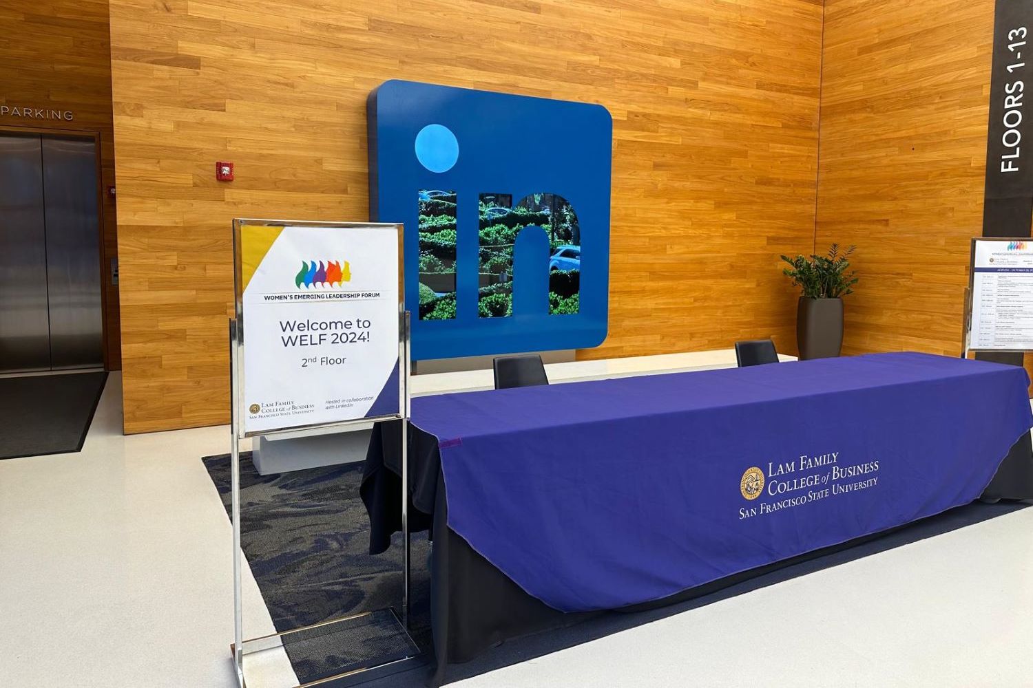 WELF 2024 -table with branded tablecloth in a lobby area, with WELF event sign and large monitor in background